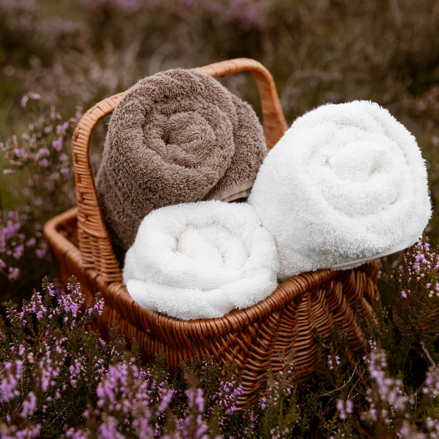 To hvide håndklæder og et brunt håndklæde i en brun, vævet kurv liggende i en eng med lyserøde blomster