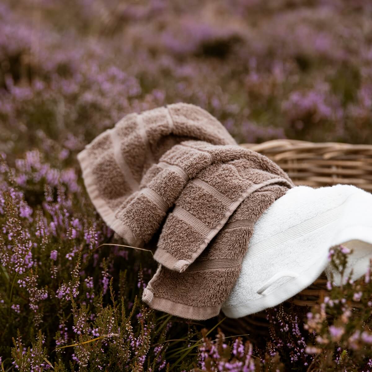 To håndklæder i brun og et håndklæde i hvidt i en brun, vævet kurv, der ligger i en eng med lyserøde blomster