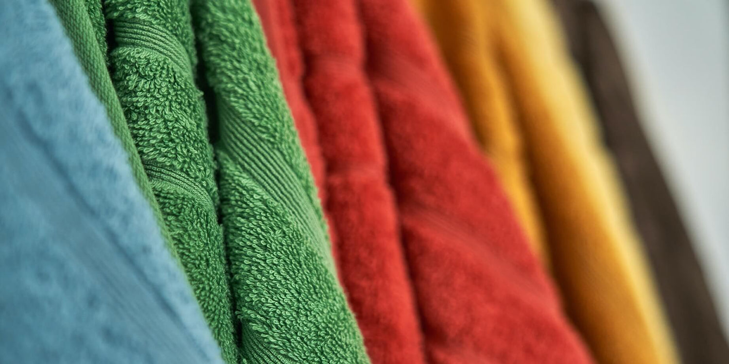 Fem håndklæder på række i farverne blå, grøn, rød, orange og brun. Teksturen er blød