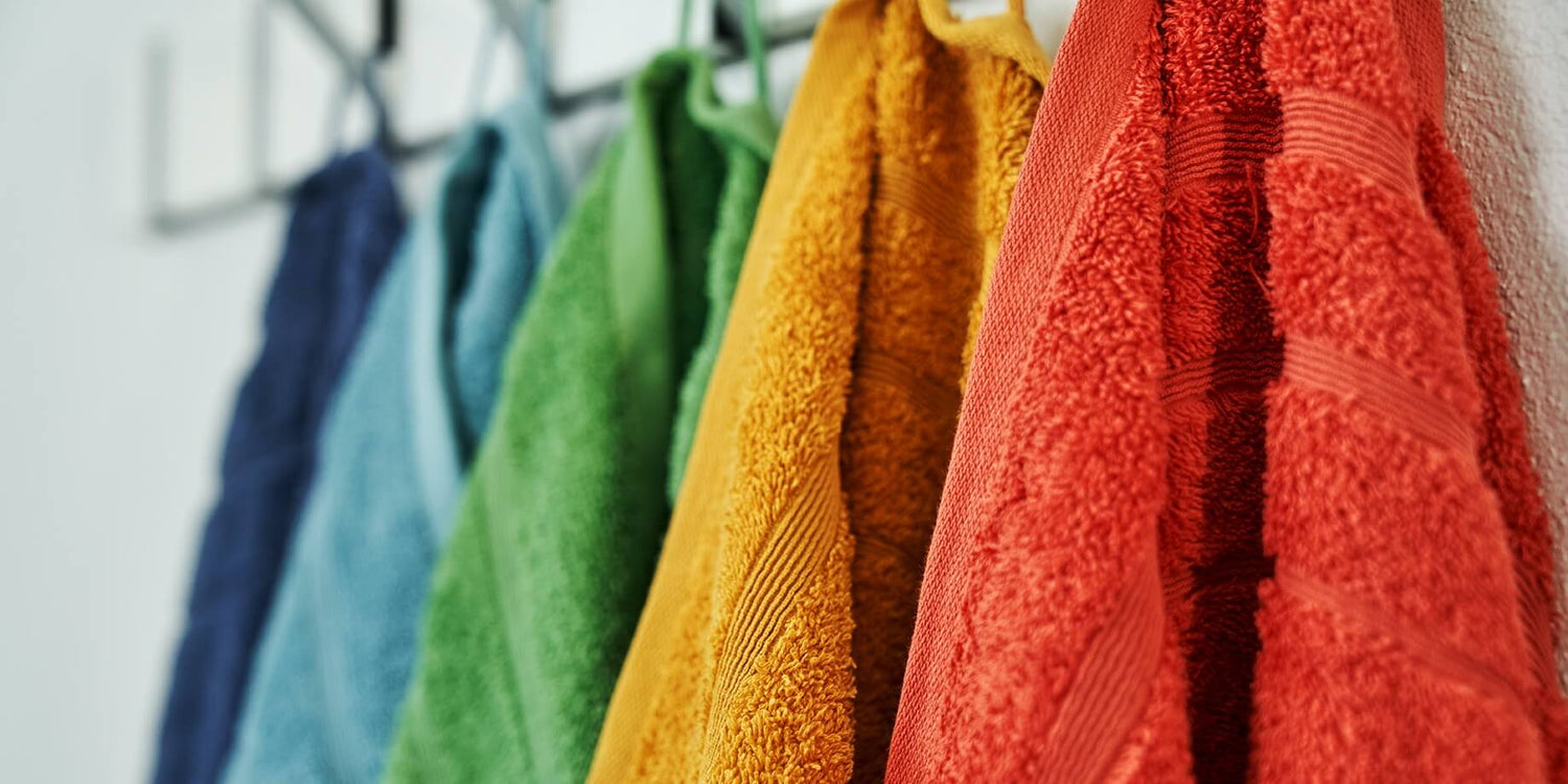 Fem håndklæder på række i farverne mørkeblå, lyseblå, grøn, orange og rød. Teksturen er blød