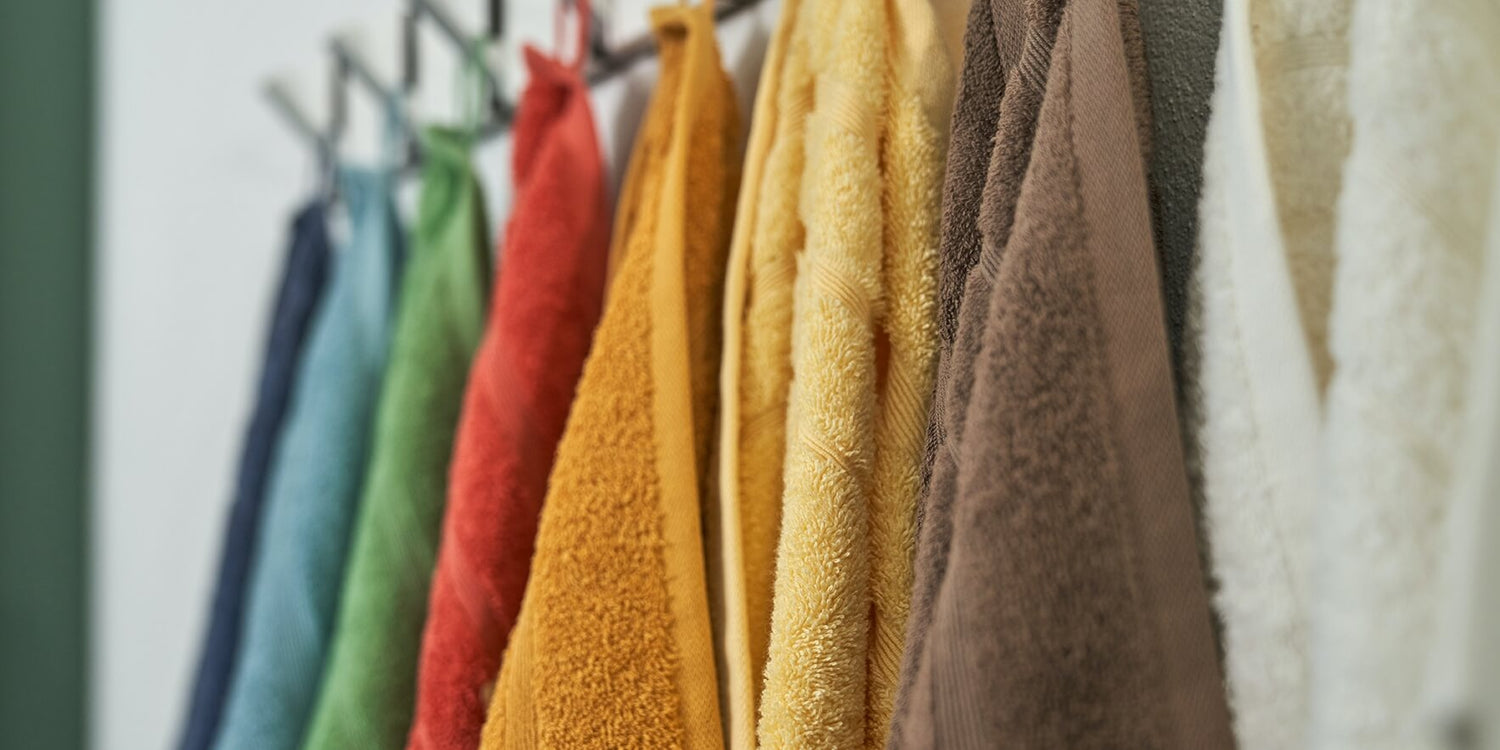 Otte håndklæder hængende på række. Farverne på håndklæderne er mørkeblå, lyseblå, grøn, rød, orange, gul, brun og hvid