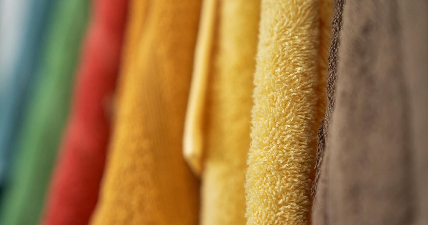 Seks håndklæder hængende på række. Farverne på håndklæderne er mørkeblå, grøn, rød, orange, gul og brun. Teksturen er blød.