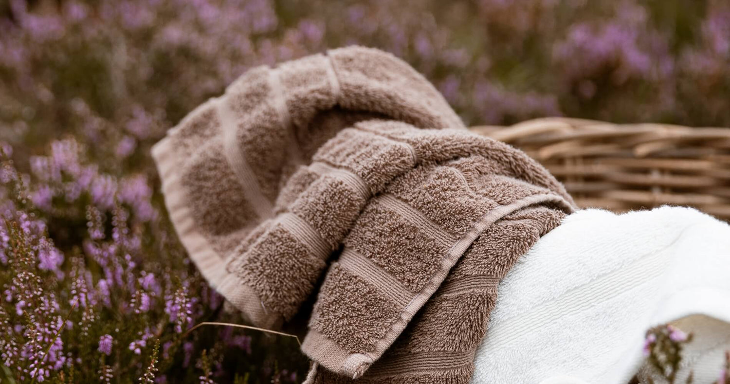 To håndklæder i brun og et håndklæde i hvidt i en brun, vævet kurv, der ligger i en eng med lyserøde blomster