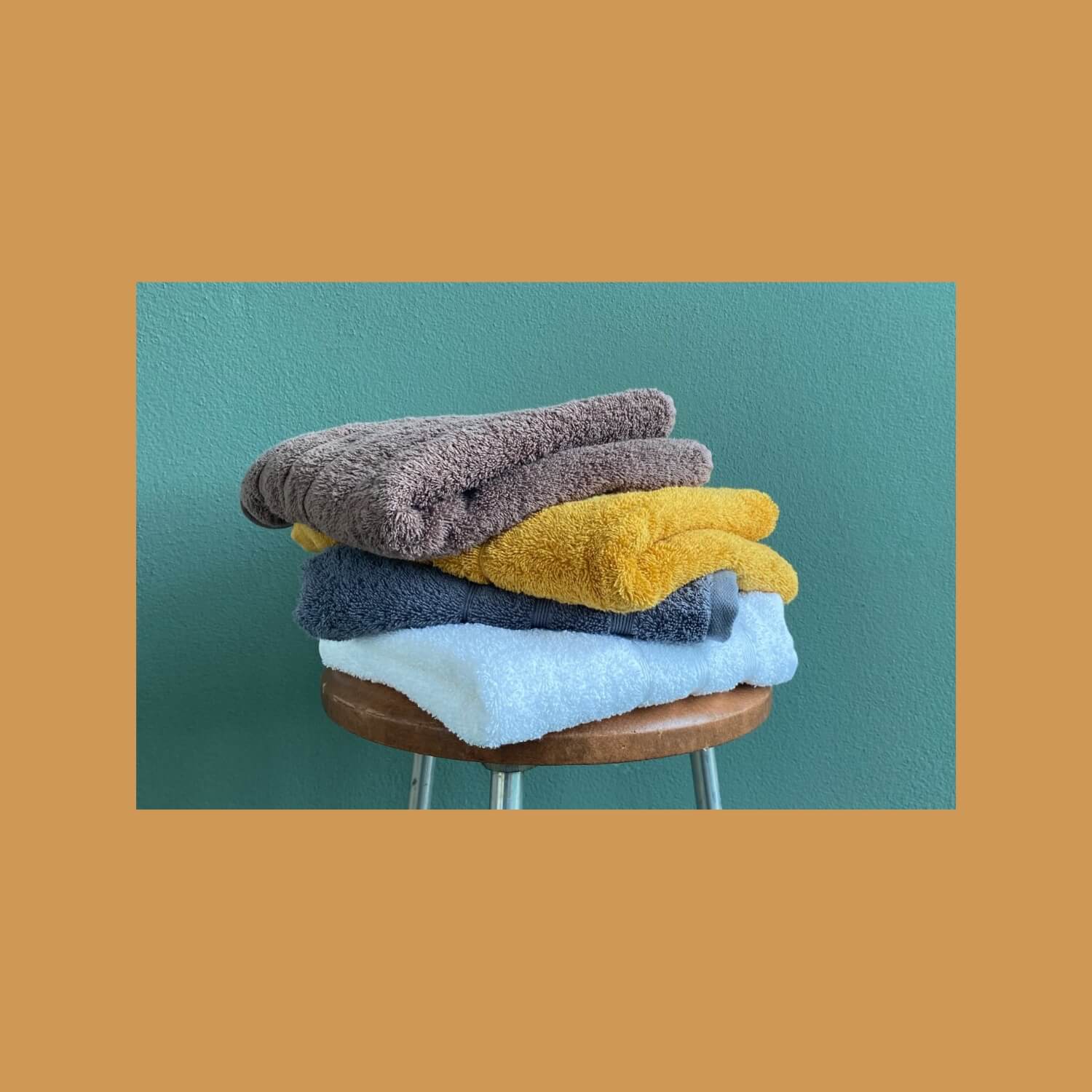 Fire håndklæder stablet oven på en skammel. Farverne er mørkebrun, karrygul, gråblå og havblå.