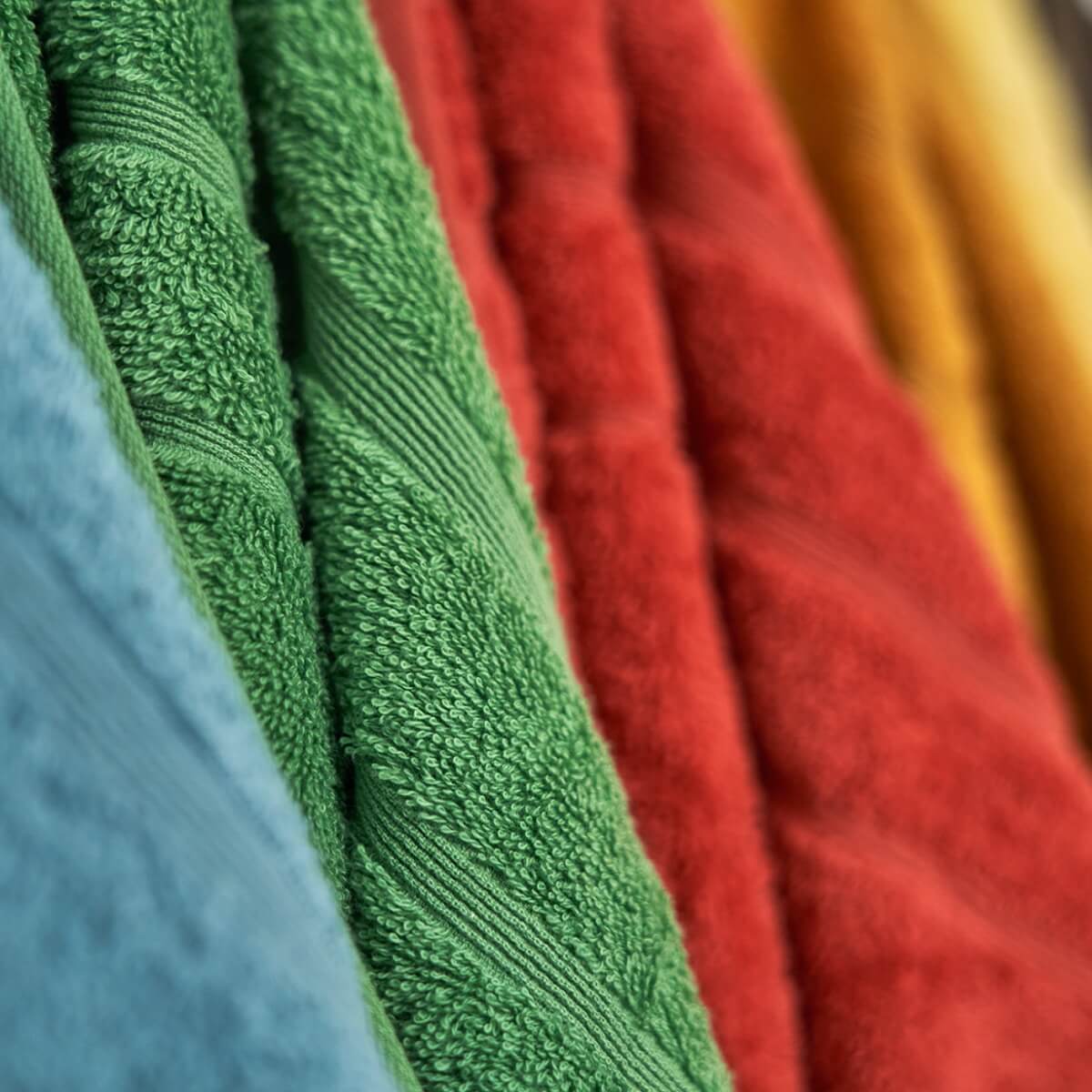 Fire håndklæder hængende på række. Farverne på håndklæderne er havblå, grøn, rød og orange. Teksturen er blød.