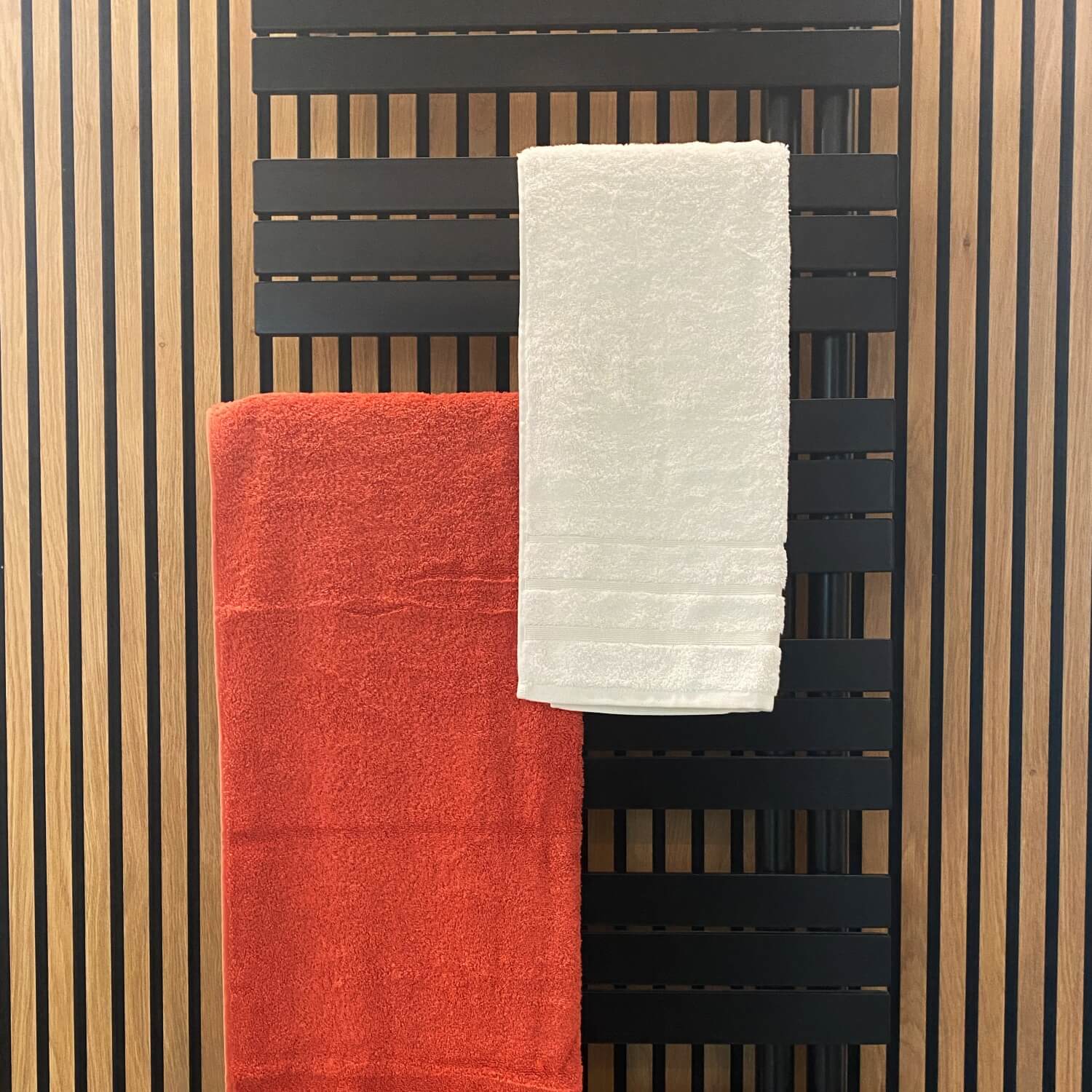 Håndklæder i rød og hvid hængende på en sort håndklædevarmer med baggrund af træpaneler 
