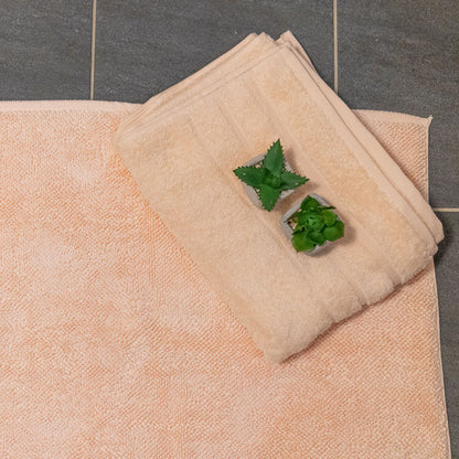 Økologisk bademåtte og håndklæde i farven melon ligger på et gråt badeværelsesgulv. 