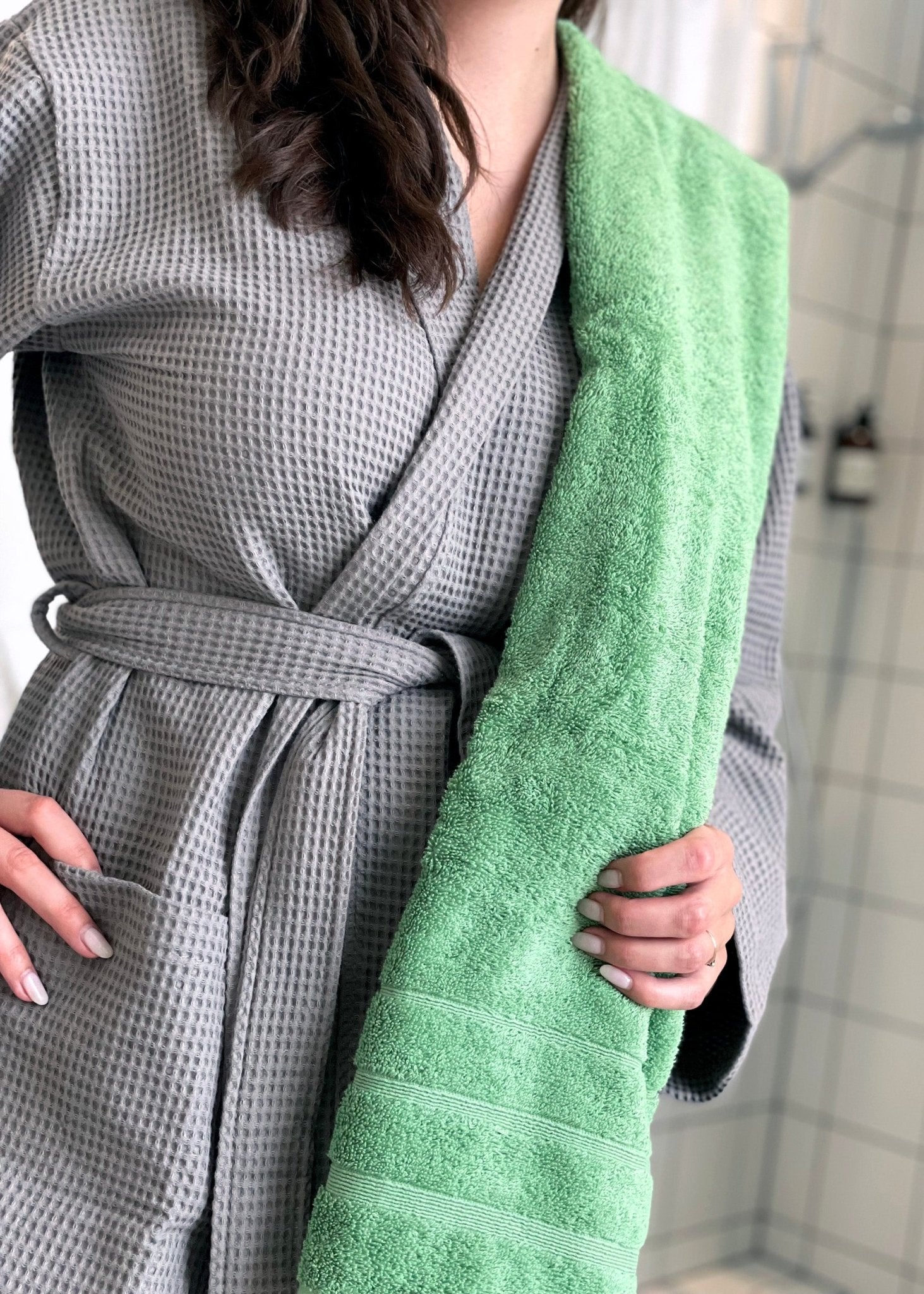Kvinde står med et grønt håndklæde over skulderen. Hun er selv iklædt en grå badekåbe. 