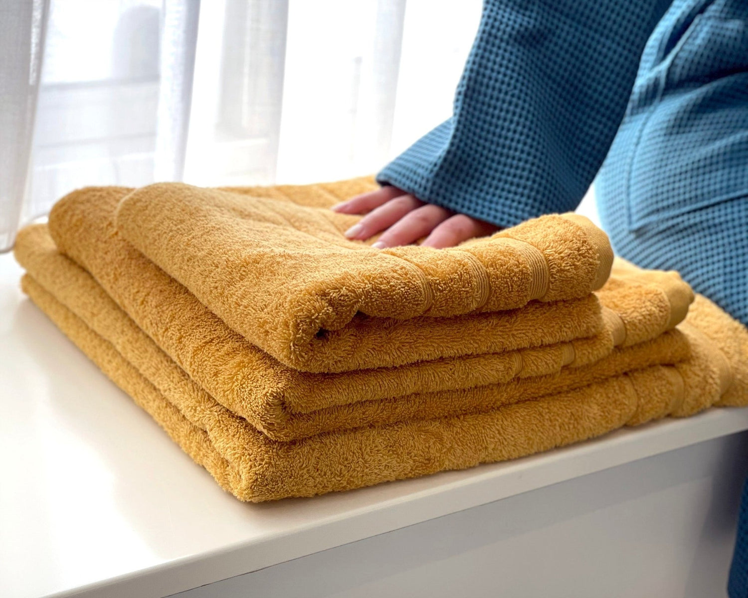 3 gule håndklæder, der ligger i en vindueskarm.En person sidder ved siden af i en blå badekåbe med hånden på håndklæderne.