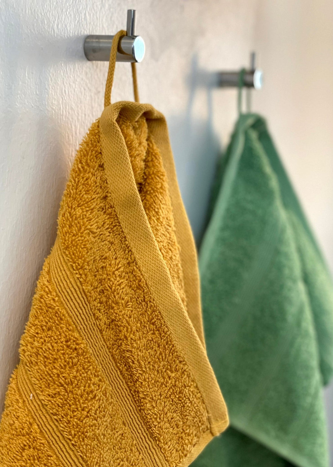 To håndklæder i farven karrygul og græsgrøn hænger på en knage inde på badeværelset.