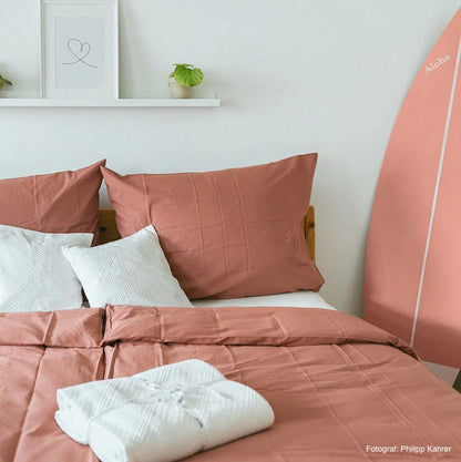 Økologisk percale sengetøj i farven rød ler. Der ligger to hvide hovedpuder og et hvidt håndklæde på den redte seng.