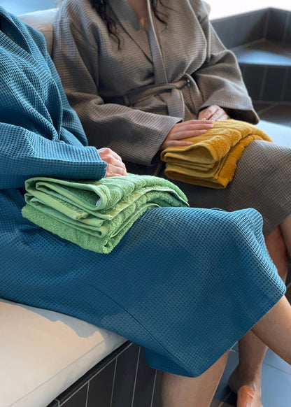 To personer sidder i en økologisk badekåbe i farven grå og blå. Den ene har et grønt håndklæde på benene.